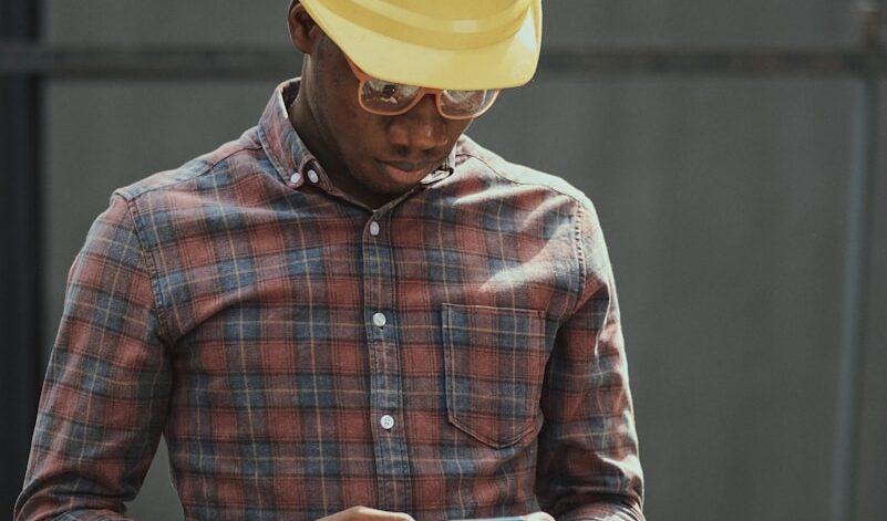 man in red blue and white plaid dress shirt wearing yellow hat holding black smartphone